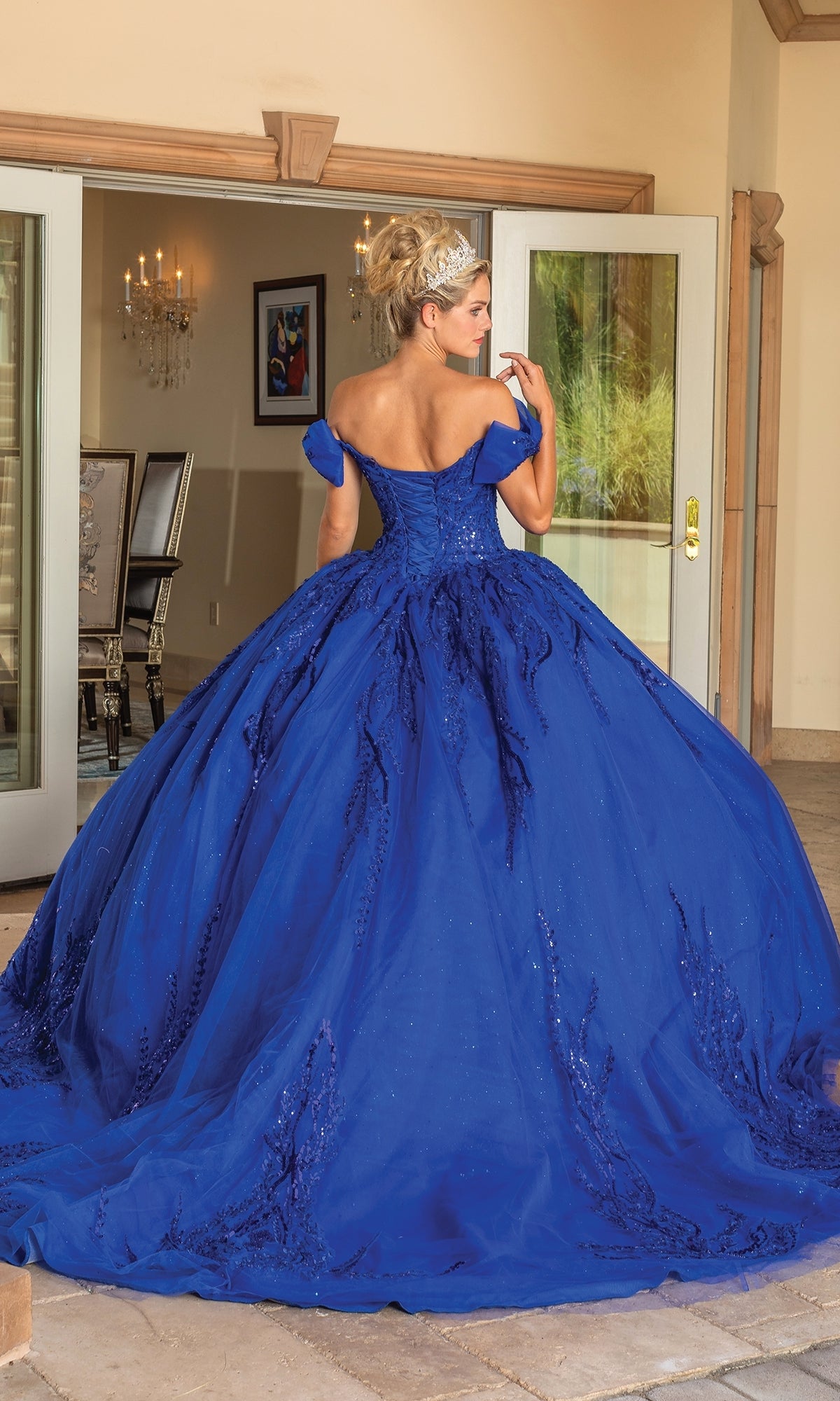 A woman with blonde hair in an updo stands near open double doors, her back to the camera, wearing the Quinceanera Dress 1780 By Dancing Queen—a royal blue, off-the-shoulder ball gown with a full skirt.